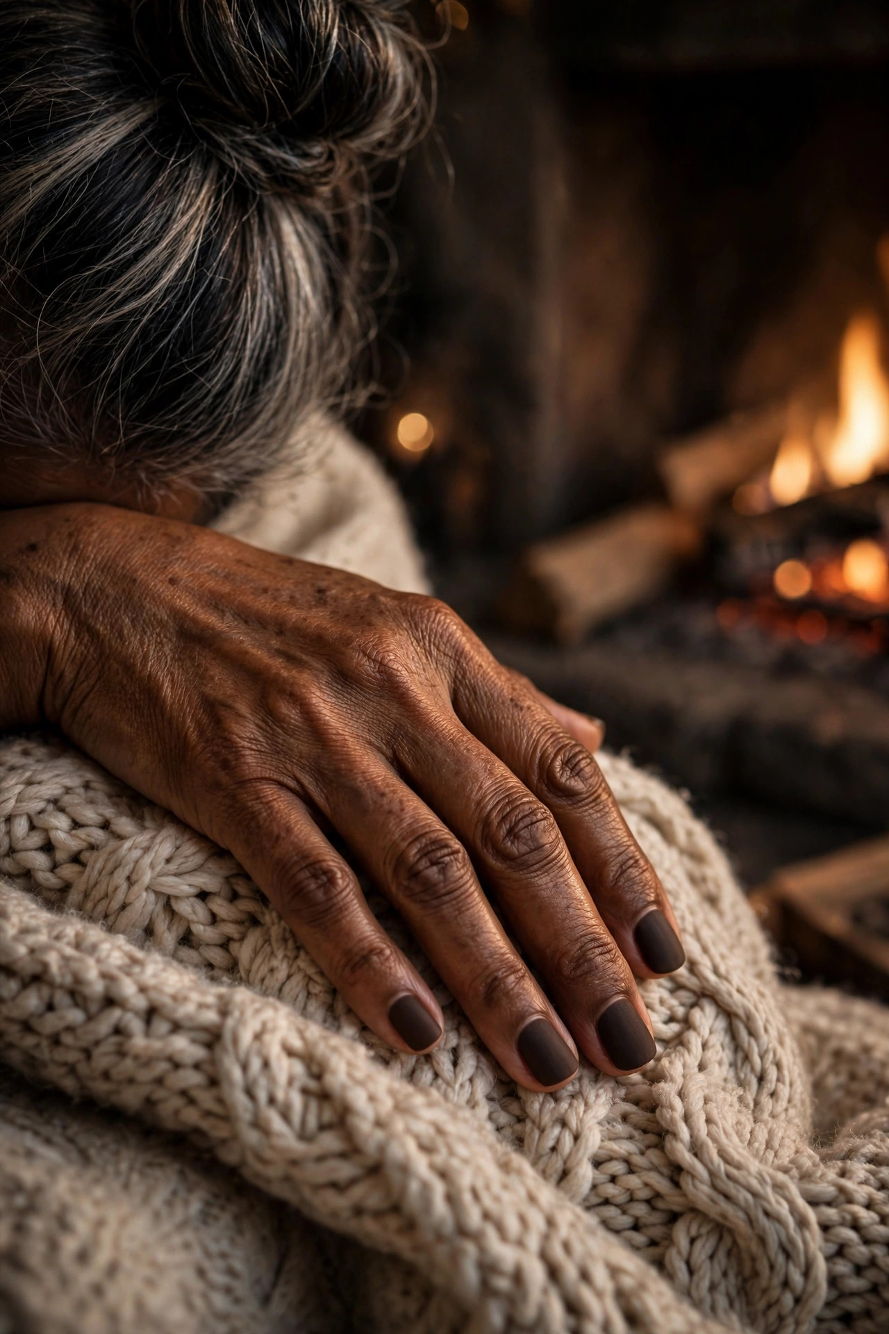 Short square matte espresso brown nails, hand resting on a chunky knit blanket near a fireplace