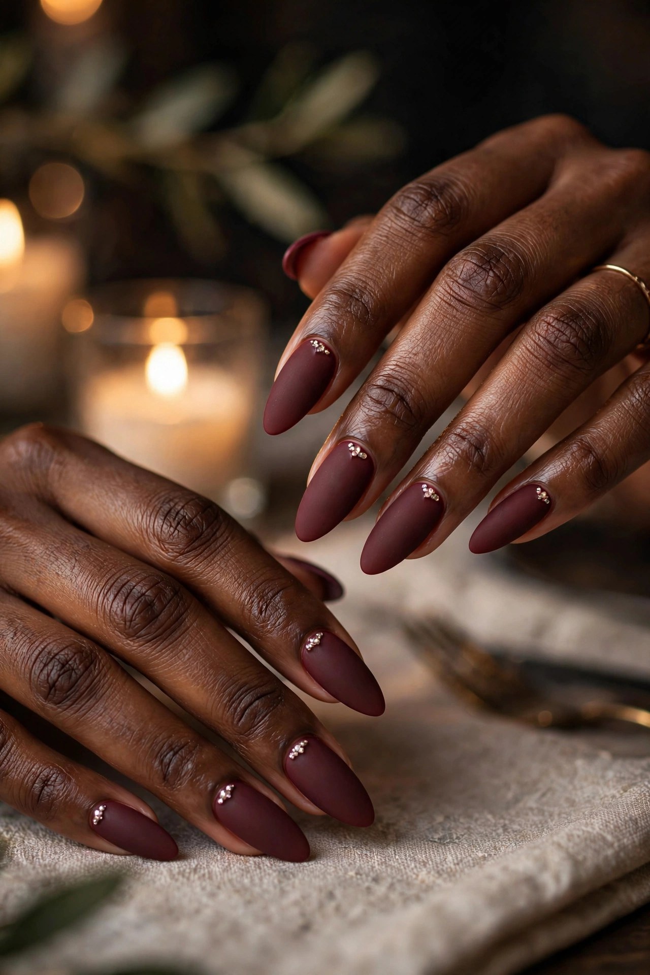 Hands with deep brown skin and long almond nails in matte burgundy with rhinestone accents